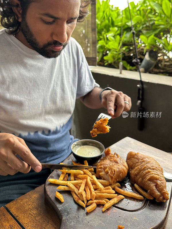 印度男子吃着金黄色的新鲜酥脆的薯条/薯条和碎鳕鱼片的图像，放在木砧板上，餐厅背景，重点在前景